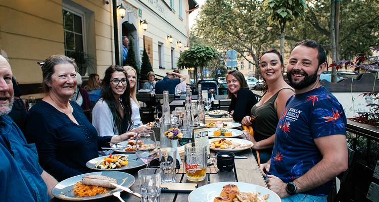 Freunde teilen ein Abendessen im Freien an einem langen Holztisch in einer grünen europäischen Straße.