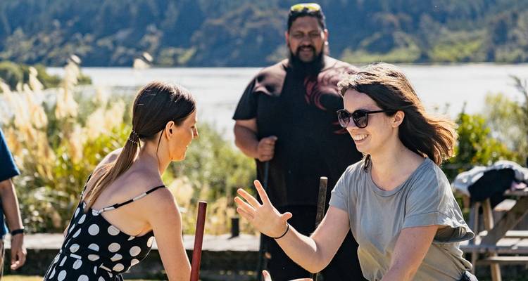 Touristen, die ein interaktives Kulturspiel an einem See mit einem Maori-Führer genießen, der zuschaut.