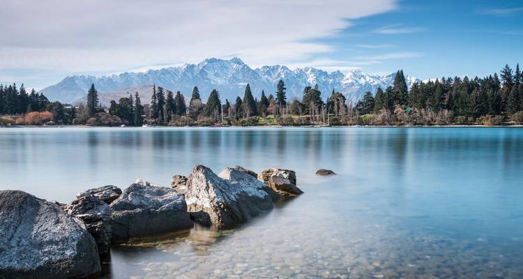 Long-exposure shot of still turquoise lake with snow-capped Remarkables mountain range beyond and rocks in the foreground.