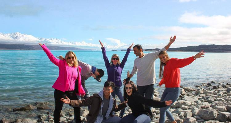 Cheerful group posing with arms raised on rocky shoreline of a bright turquoise lake backed by snowy peaks.