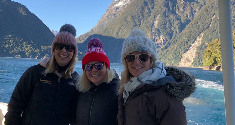 Three women in winter hats smiling on the deck of a boat cruising through a dramatic fjord.