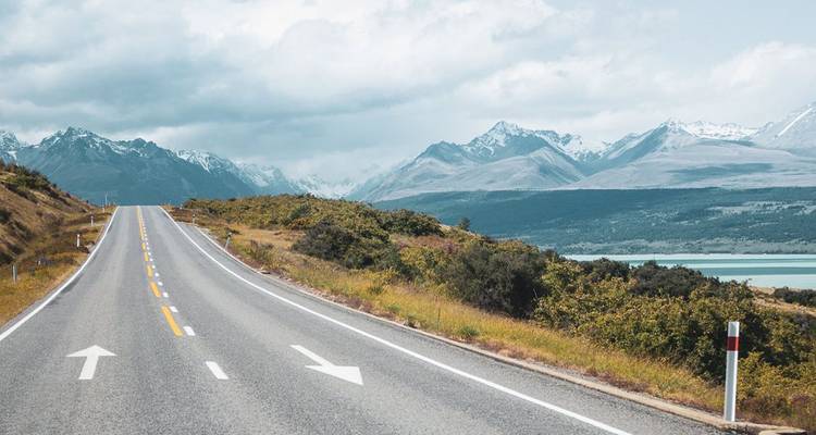 Open highway with white arrows stretching toward distant snow-capped mountains and a turquoise lake.