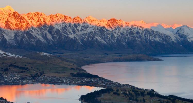 Golden sunrise light illuminating snowy peaks above a mirror-like lake and small town below.