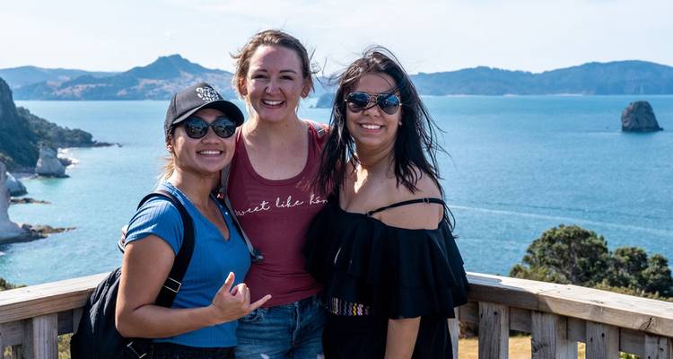 Trois amis sourient sur une plateforme d'observation en bois surplombant une baie bleu vif avec des formations rocheuses et des collines boisées.