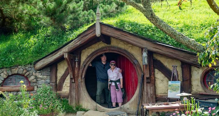 Un couple se tient dans l'embrasure ronde d'un terrier de hobbit à Hobbiton, entouré de fleurs et d'herbe luxuriante.