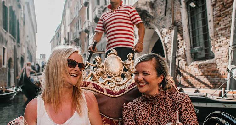 Deux femmes rient dans une gondole décorée tandis qu'un gondolier vêtu d'une chemise rayée rame dans un étroit canal de Venise.
