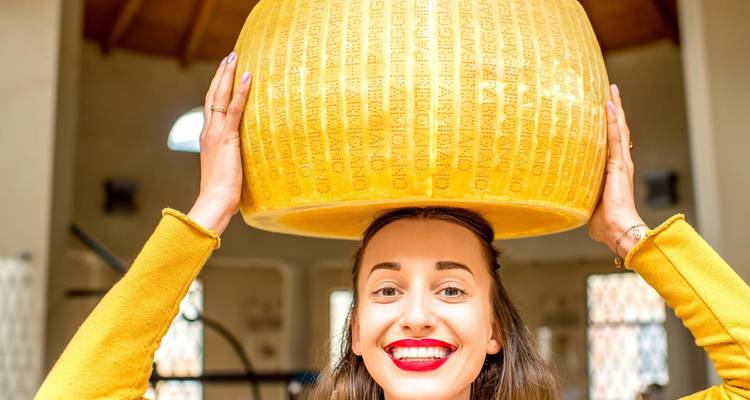 Smiling woman balancing a giant wheel of Parmigiano-Reggiano cheese on her head in a dairy