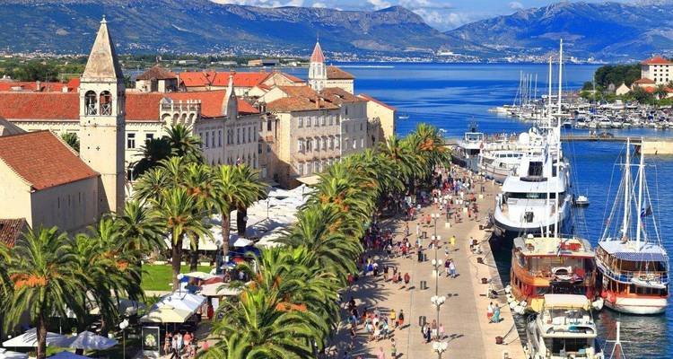 Lively seaside promenade in Trogir lined with palm trees, yachts and historic stone buildings.