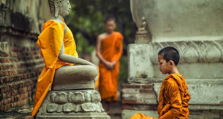 Un jeune moine novice s'agenouille en méditation devant une statue de Bouddha en pierre drapée de robes safran dans une cour ancienne.