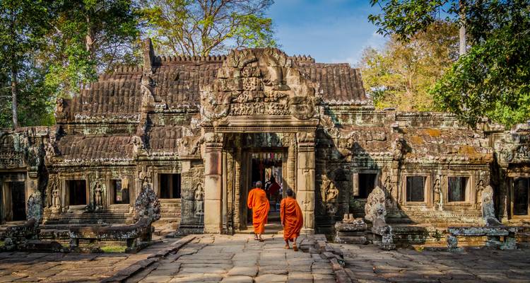 Twee monnikken in oranje gewaden die een versleten ingang van een Angkor-tempel binnenlopen, beschaduwd door hoge bomen.