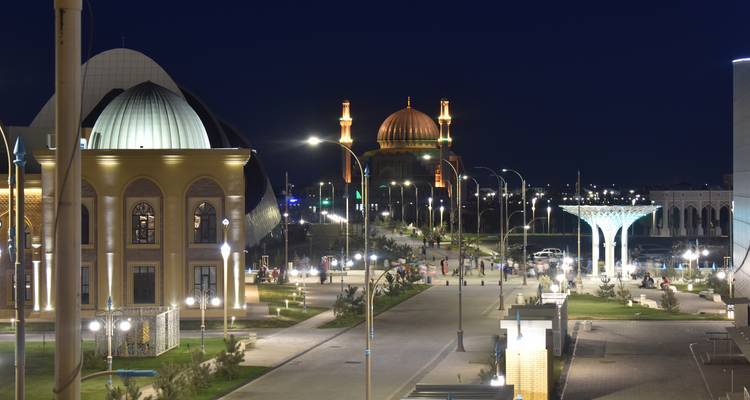 Nachtansicht einer Boulevardstraße in Turkestan mit beleuchteten modernen Gebäuden, einer Moschee-Kuppel und spazierenden Besuchern.