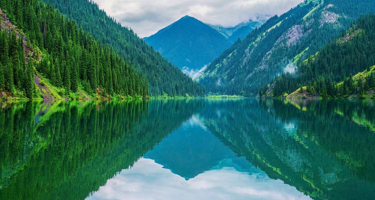 Kristallklarer Alpensee, der schneebedeckte Berge unter einem teilweise bewölkten Himmel in Kasachstan widerspiegelt.