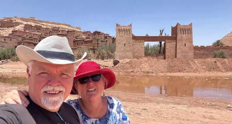 Selfie of couple in hats with earthen ramparts and gate of Ait Benhaddou in background.