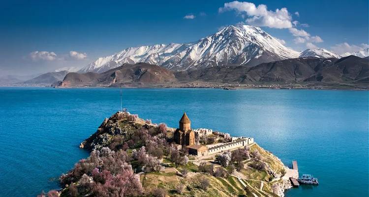 Island with historic stone church and fortress surrounded by blooming trees on turquoise Lake Van with snow-capped mountain backdrop.