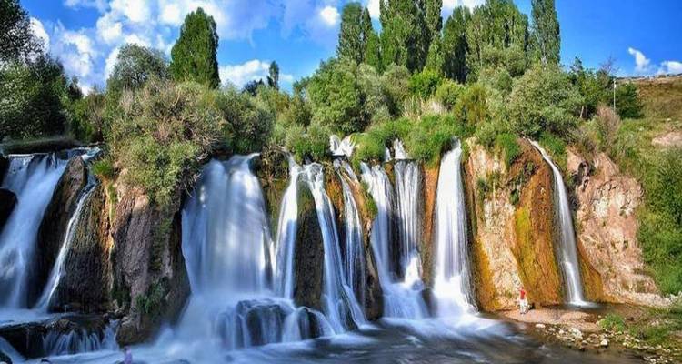 Broad multi-cascade waterfall surrounded by lush greenery under bright blue sky.