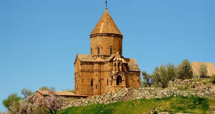 Brown-stone Akdamar Church surrounded by spring blossoms on green hillside under clear sky.