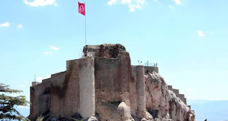 Stone fortress atop rocky hill flying Turkish flag against bright sky.