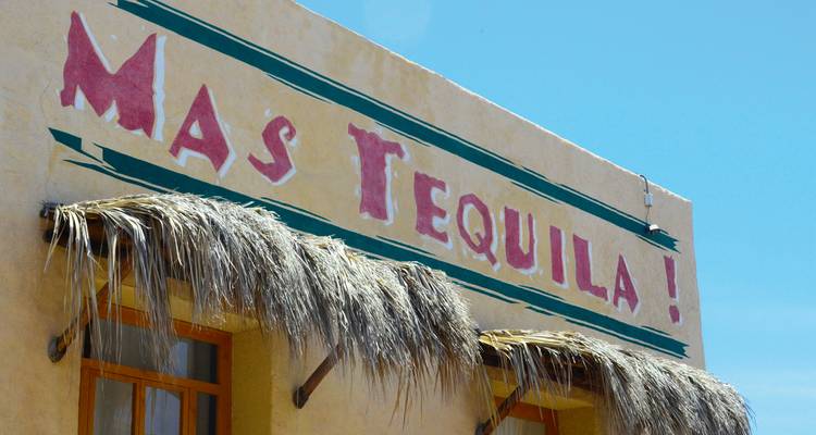 Fachada de edificio de adobe con un letrero pintado a mano que dice 'Mas Tequila!' bajo un cielo azul brillante