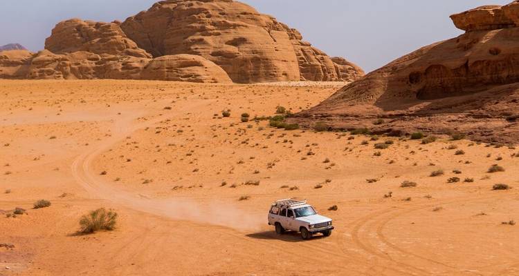Ein 4x4-Fahrzeug fährt über weite orangefarbene Sandebenen unter Sandsteinformationen im Wadi Rum.