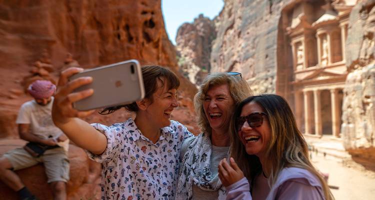 Drei Frauen lachen, während sie ein Selfie vor der ikonischen geschnitzten Fassade von Petras Schatzkammer in einer engen Sandsteinkluft machen.