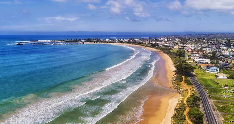 An aerial view of a sweeping crescent beach, turquoise sea and coastal town backed by rolling green hills.
