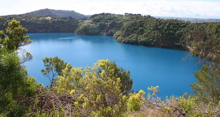A striking deep-blue crater lake is surrounded by lush green vegetation and forested ridges.
