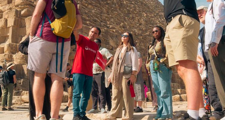 Un guide touristique en chemise rouge gesticule tandis qu'un petit groupe de voyageurs se tient à côté des énormes blocs de calcaire d'une pyramide sous un ciel bleu.