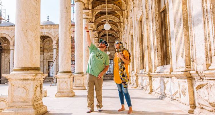 Deux voyageurs admirent des colonnes de marbre ornées et des arches à l'intérieur d'une cour de mosquée historique baignée de lumière vive.