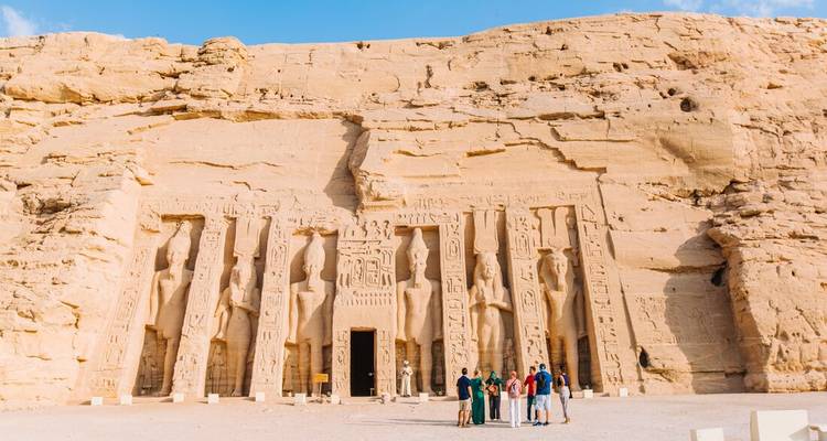 Des statues colossales taillées dans la roche font face à la façade de grès d'Abou Simbel tandis qu'un petit groupe de touristes se rassemble à l'entrée sous un ciel bleu éclatant.
