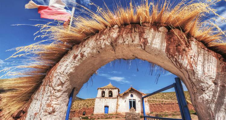 Rustic adobe arch crowned with golden straw framing a small Andean church under a bright blue sky.