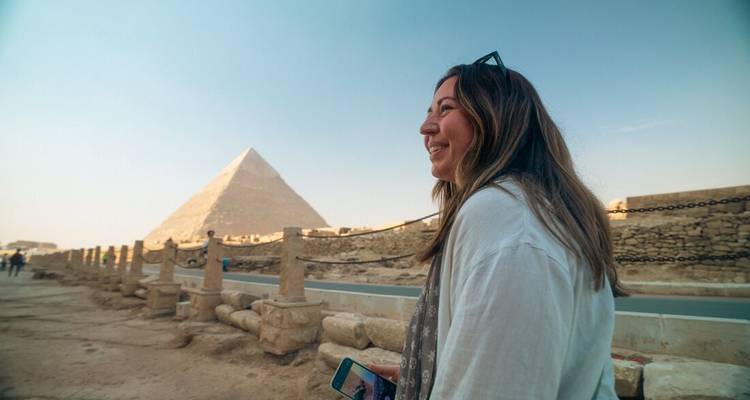 Smiling traveller holding a phone with the Pyramid of Giza softly lit in the background against a clear blue sky.