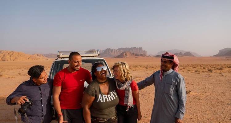 Laughing friends gather beside a 4x4 in the vast red desert of Wadi Rum with rugged sandstone peaks behind.
