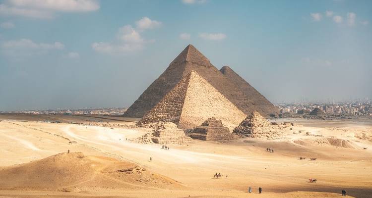 Panoramic vista of the three Pyramids of Giza rising above sweeping golden sands beneath a bright blue sky.