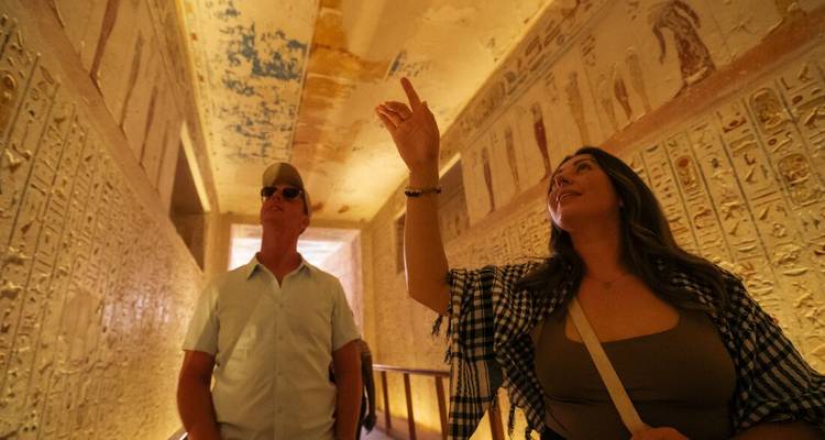 Two travellers study vivid wall paintings and ceilings inside an elaborately decorated pharaonic tomb.