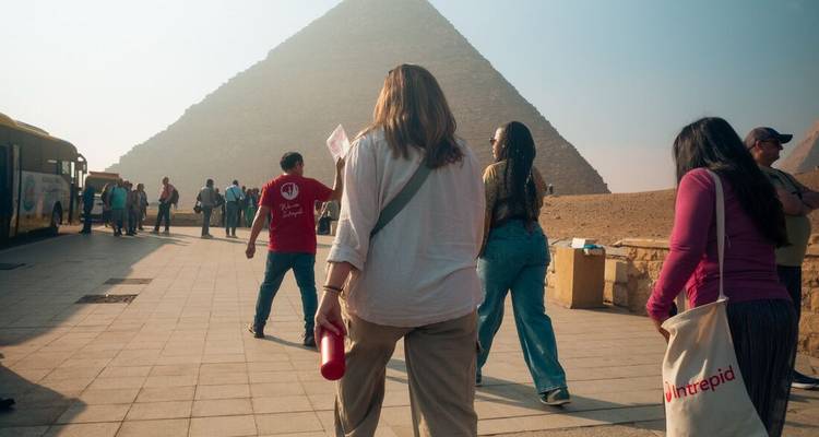 Travellers walk toward a pyramid complex on a hazy morning carrying water bottles and tour materials.