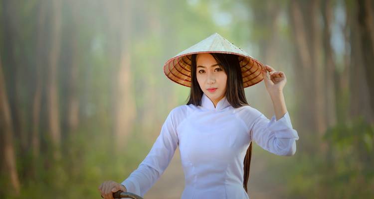 Elegant Vietnamese woman in white ao dai adjusting her conical hat on a tree-lined path bathed in soft morning light.