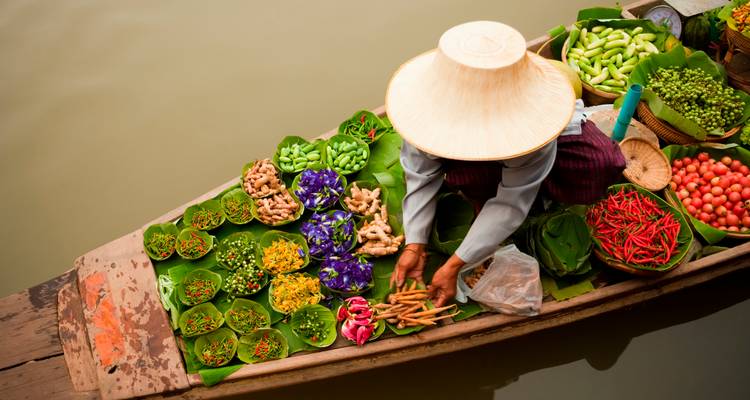 Vendor kneeling in a wooden boat arranging an array of vibrant fruits, vegetables and flowers on banana leaves.