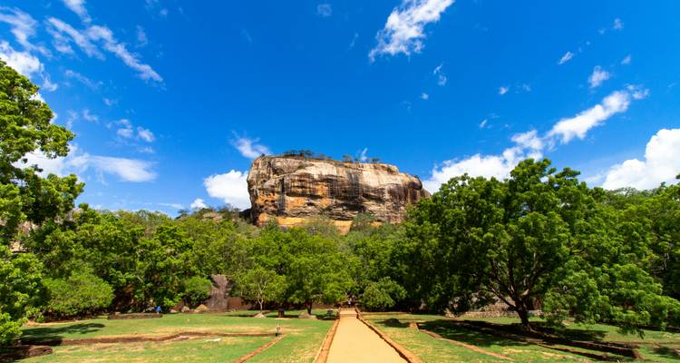 Wide pathway leading to the imposing Sigiriya rock fortress under a brilliant blue sky and lush greenery.