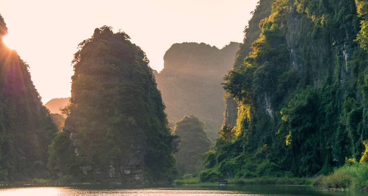 Sunset light illuminating steep karst cliffs rising from a calm river in a lush Vietnamese valley.