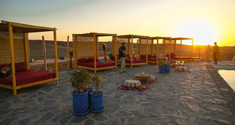 Desert camp terrace at sunset with bamboo cabanas, loungers, low tables and guests enjoying the golden horizon by a pool.