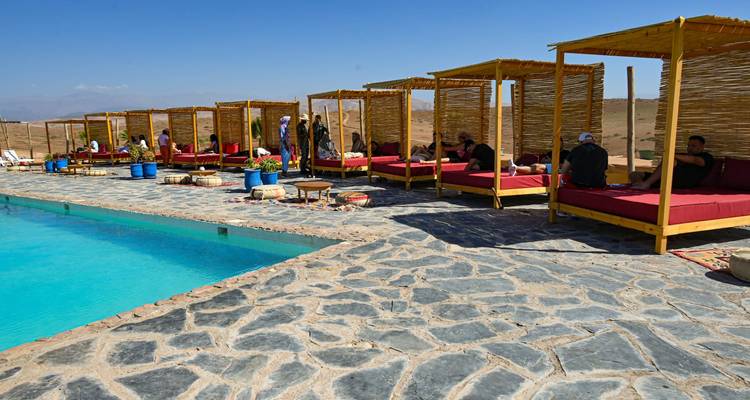 Daytime view of a stone-paved pool deck in the desert lined with bamboo cabanas where guests lounge by turquoise water.