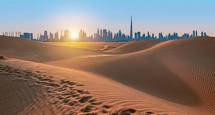 Rollende orange Wüstendünen mit der Skyline von Dubai, die sich gegen eine untergehende Sonne abzeichnet.
