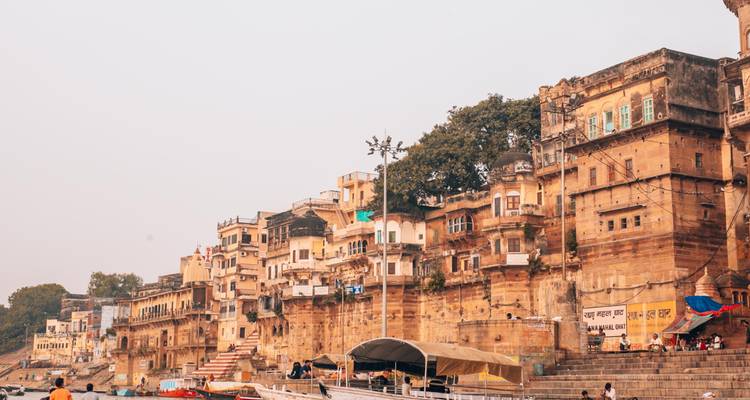 Historic sandstone ghats and houses line the Ganges riverbank in hazy Varanasi light.