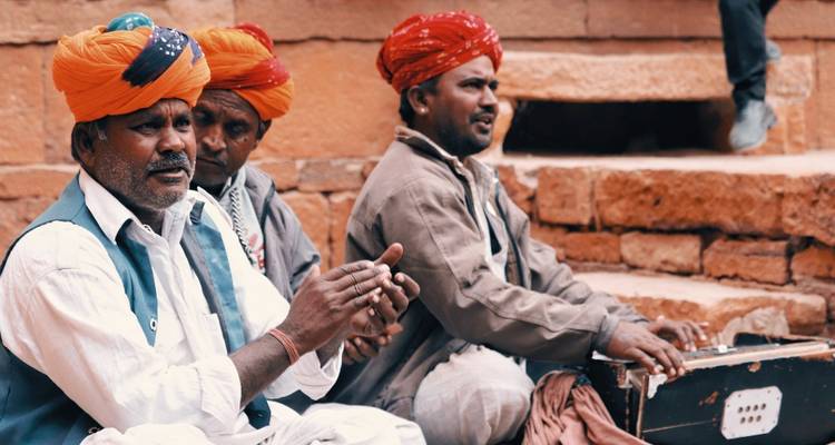 Traditional Rajasthani musicians wearing bright turbans clap and play instruments against red stone walls.