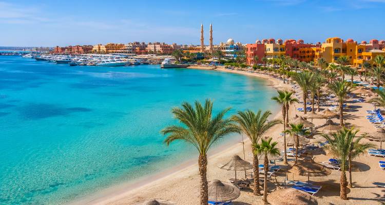 Tropical Red Sea beach lined with palms, umbrellas and colorful Hurghada resorts beside turquoise water.