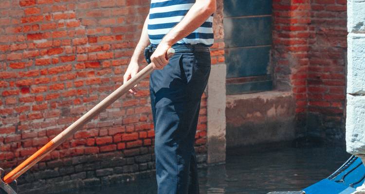 Gondolier traditionnel en chemise rayée debout avec une rame dans un étroit canal vénitien