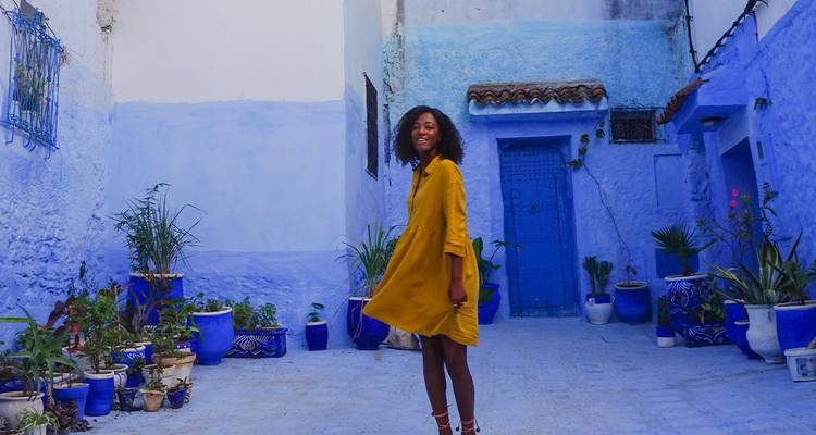 Femme souriante vêtue d'une robe jaune posant dans une ruelle bleu vif de Chefchaouen.