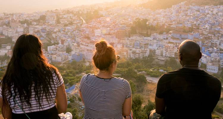 Trois voyageurs sont assis sur une crête surplombant les maisons aux couleurs pastel de Chefchaouen baignées de lumière dorée.