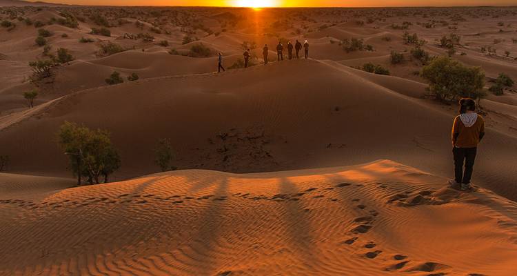 Les voyageurs marchent le long de la crête des dunes ondulantes du Sahara qui rougeoient d'orange sous le soleil couchant.