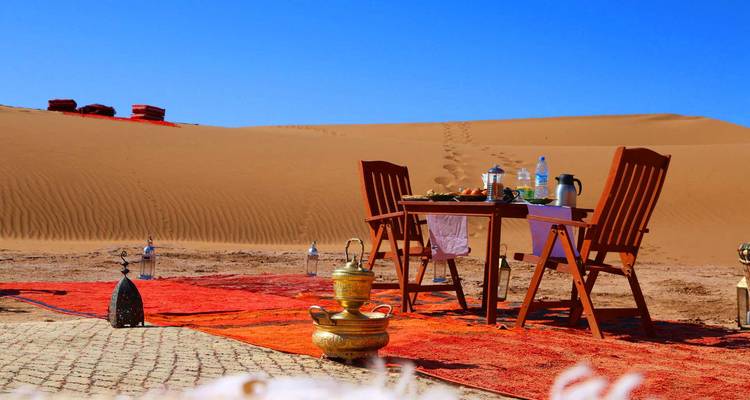 Table de petit-déjeuner dressée sur des tapis colorés sous d'imposantes dunes de sable